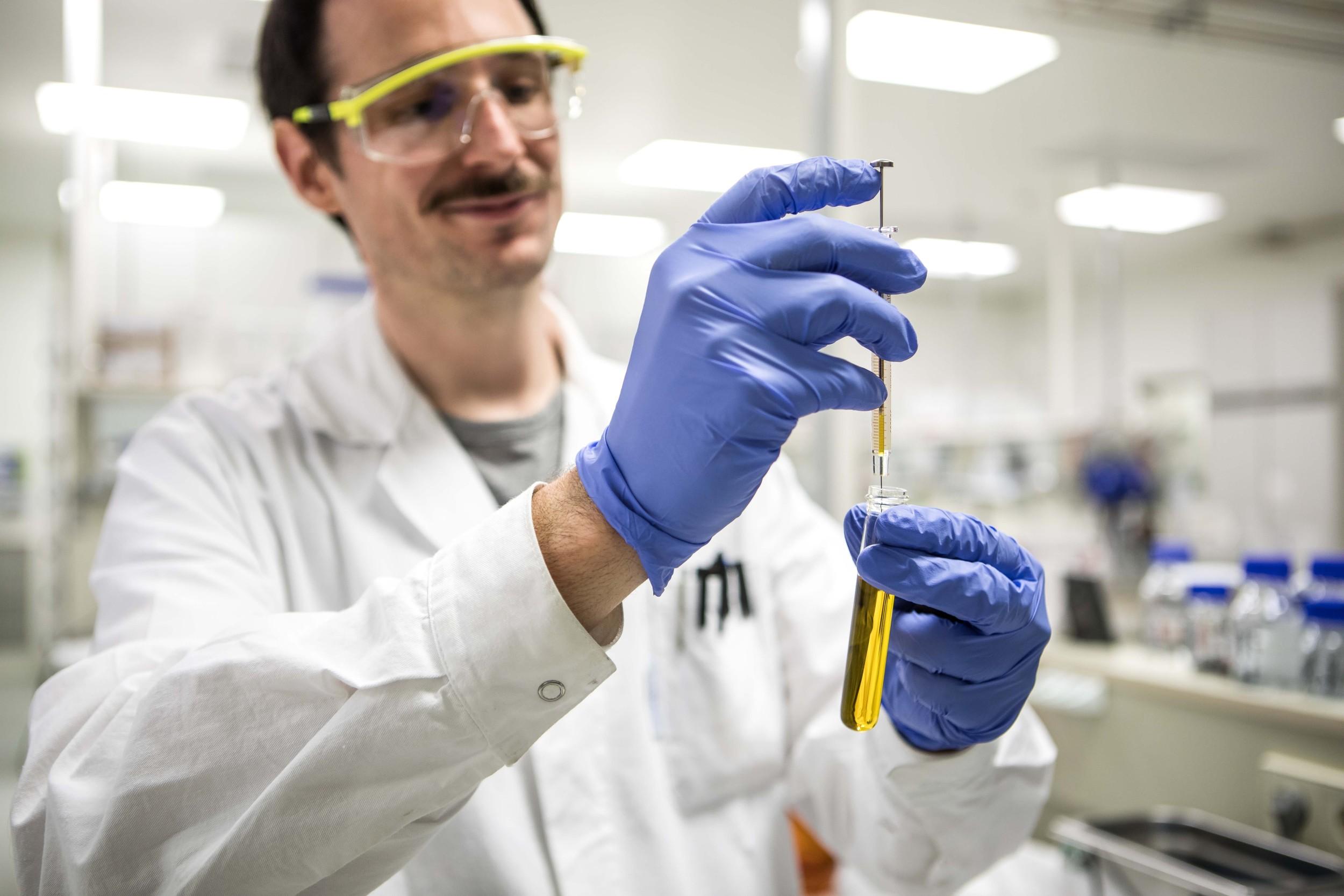 Laboratory technician wearing protective gloves and goggles, carefully transferring a yellow liquid into a test tube using a pipette in a pharmaceutical or scientific lab.